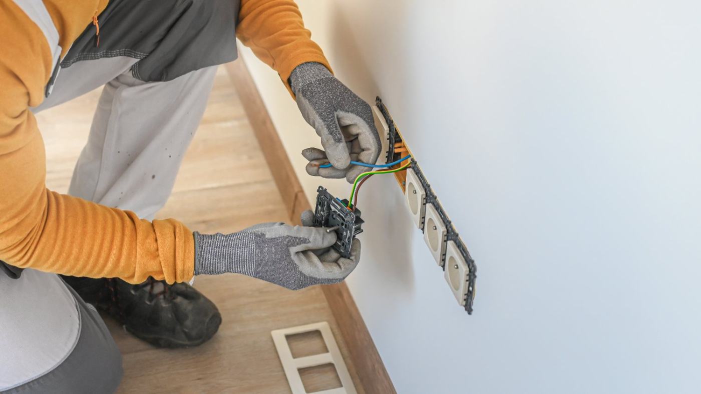 Electrician in orange sleeves installing wiring and outlet on white wall